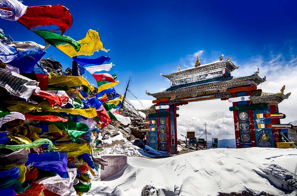 Colorful prayer flags and the Welcome to Tawang gateway at a snow-covered mountain pass in Arunachal Pradesh, Northeast India.
